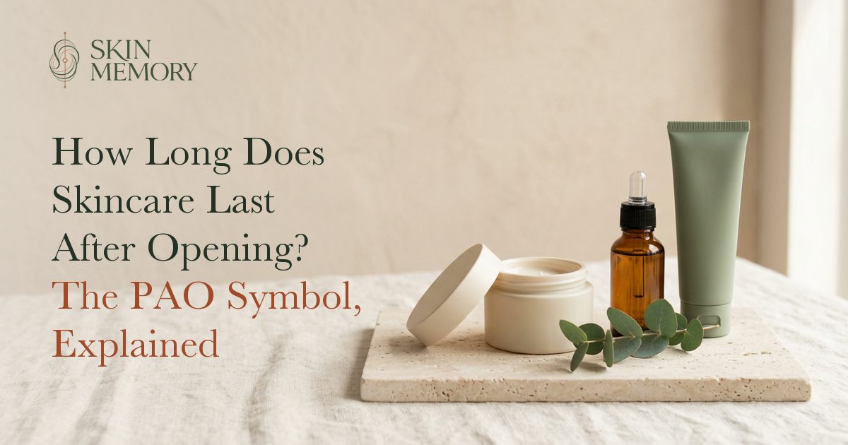 An opened jar of face cream with its lid beside it, an amber dropper bottle and a sage tube on a stone slab, illustrating how long skincare lasts after opening
